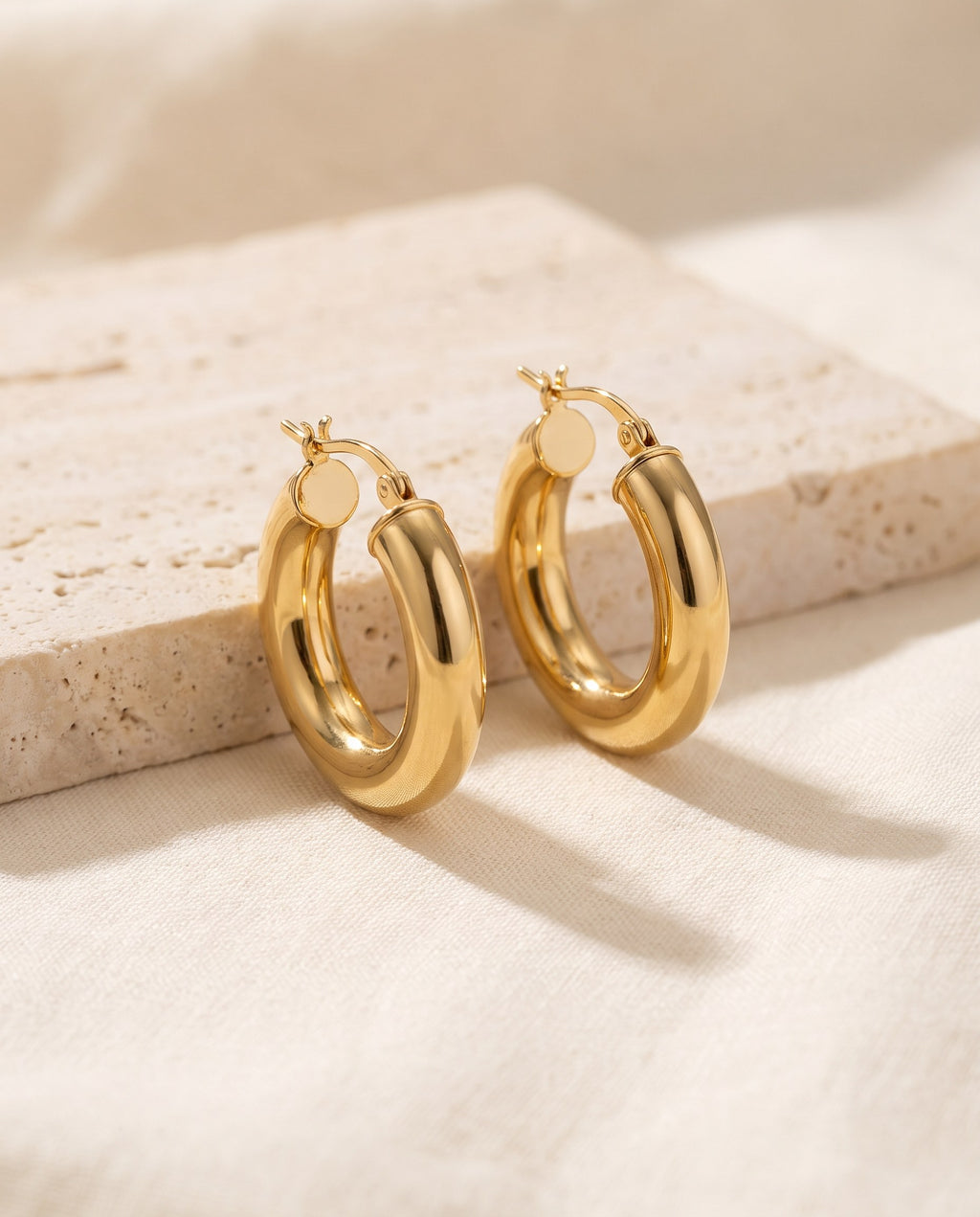 Pair of chunky gold-plated Dome Hoop Earrings, 20 mm, on a travertine block over ivory linen