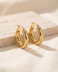 Pair of chunky gold-plated Dome Hoop Earrings, 20 mm, on a travertine block over ivory linen