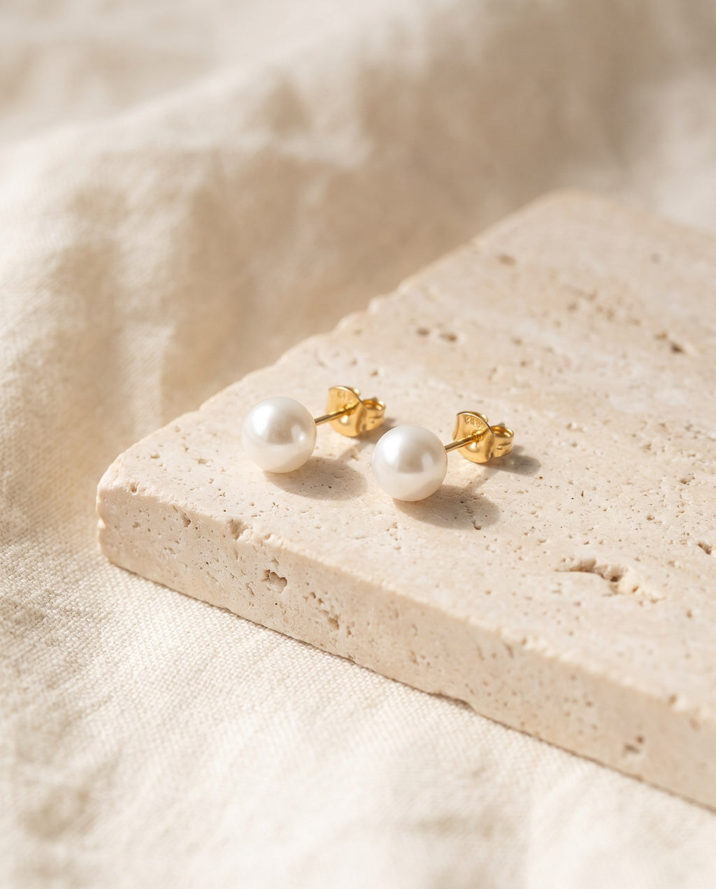 Pair of white Glass Pearl Stud Earrings with gold-plated posts on a travertine block over ivory linen