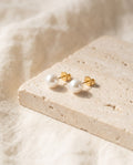 Pair of white Glass Pearl Stud Earrings with gold-plated posts on a travertine block over ivory linen