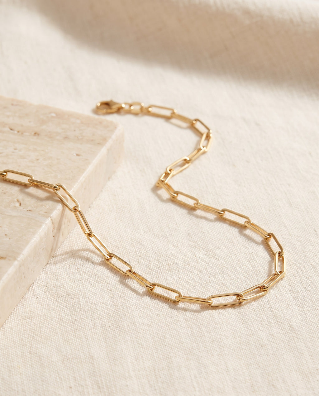 Paperclip Chain Necklace in 18k gold-plated stainless steel on ivory linen beside a travertine block