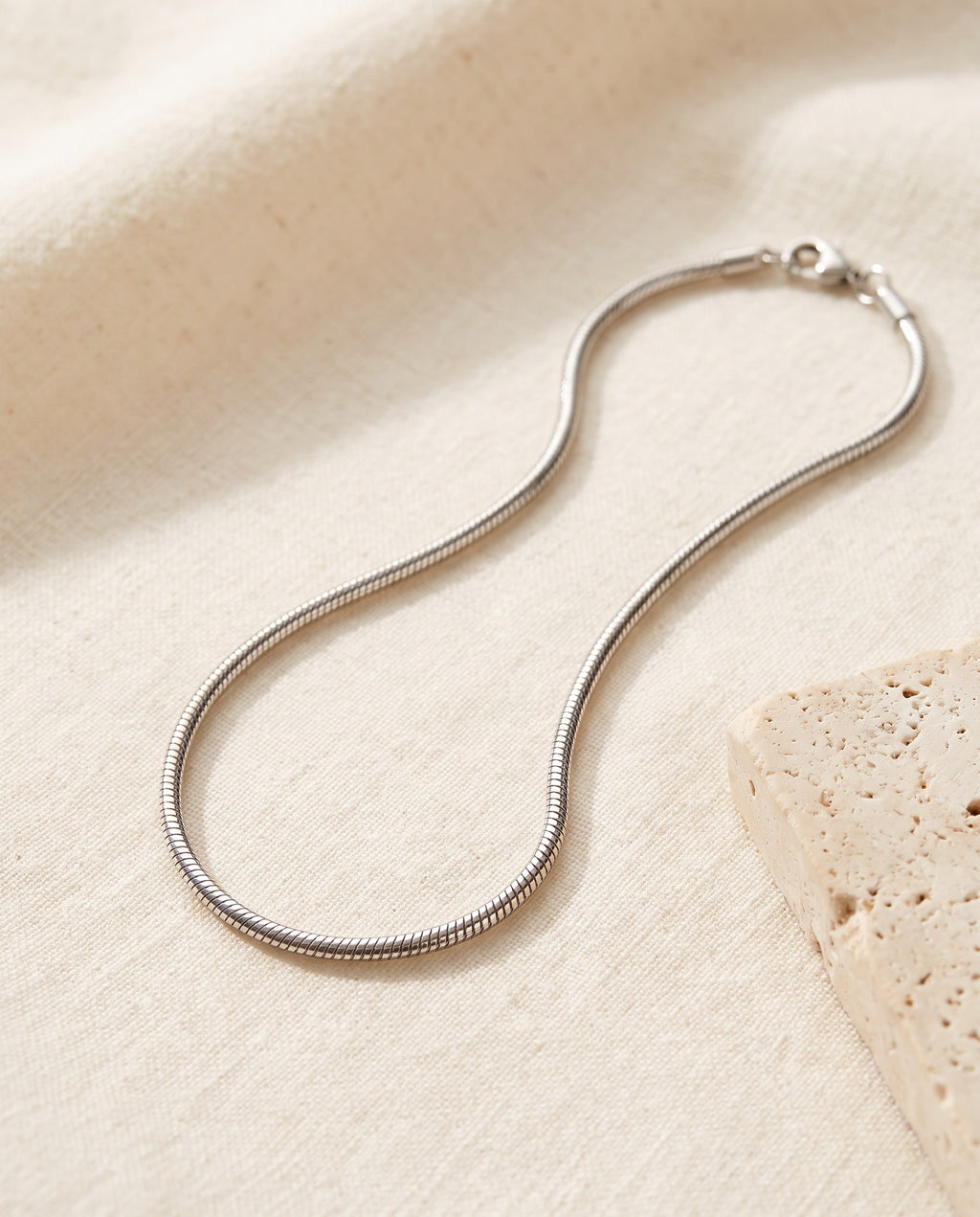 Silver-tone Snake Chain Necklace in polished stainless steel on ivory linen beside a travertine block