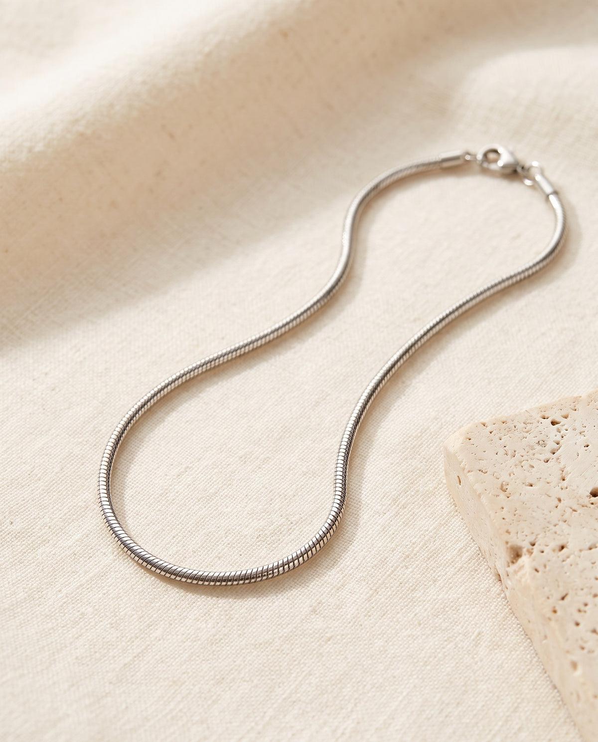 Silver-tone Snake Chain Necklace in polished stainless steel on ivory linen beside a travertine block